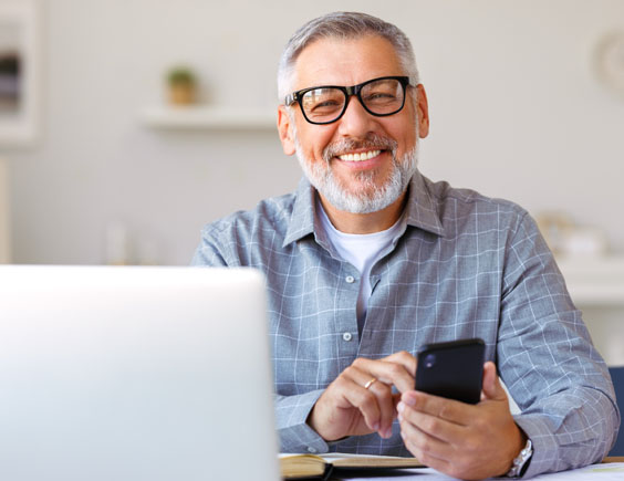 Hombre mayor sonriendo y consultando su teléfono móvil y el ordenador